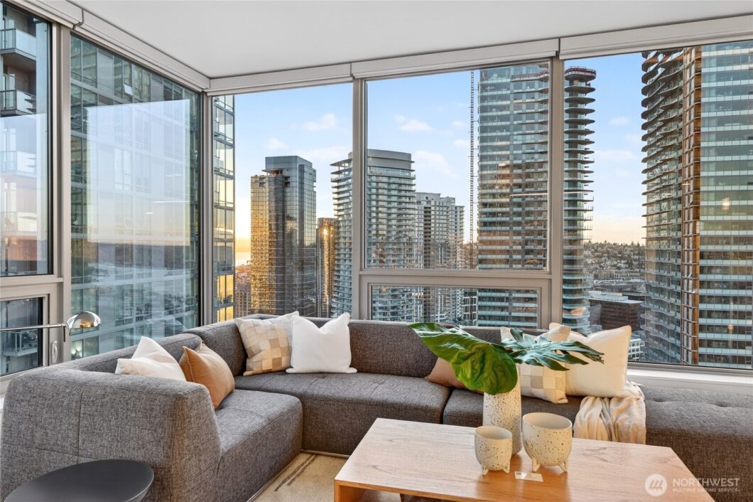 A grey sectional sofa sits in a living room with large windows overlooking city buildings.