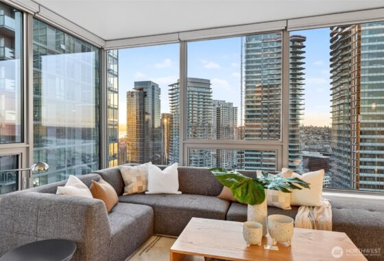 A grey sectional sofa sits in a living room with large windows overlooking city buildings.