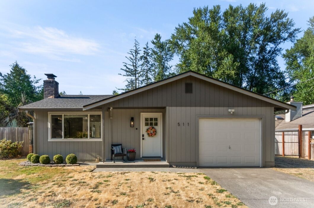 A beige single-story house with a white front door, attached garage, and tall trees behind it.