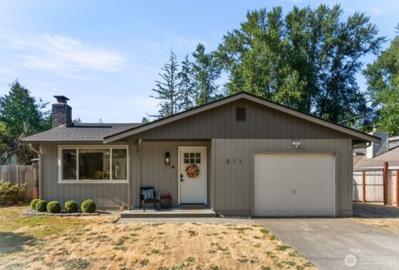 A beige single-story house with a white front door, attached garage, and tall trees behind it.
