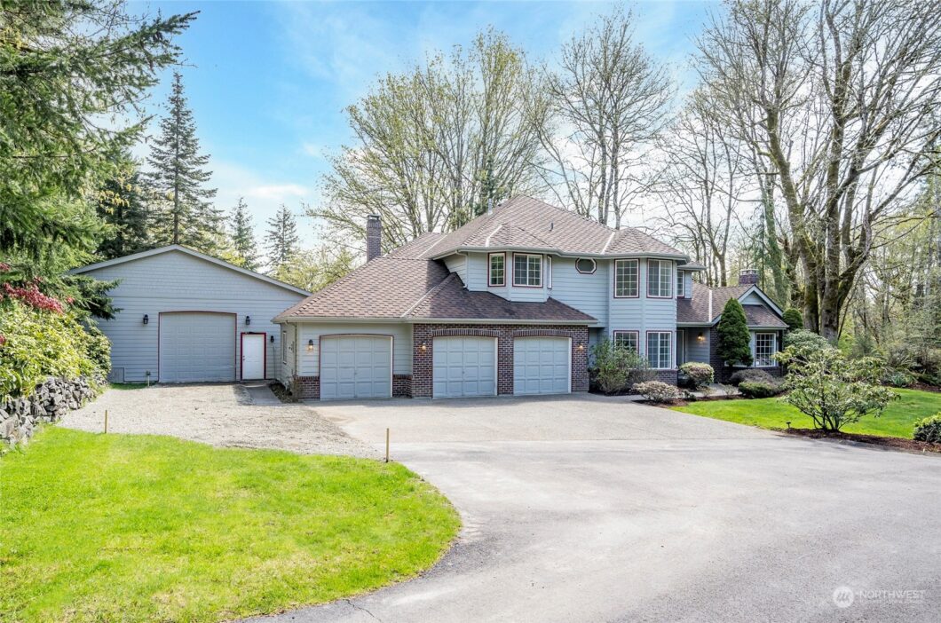 A two-story house with a three-car garage and driveway surrounded by trees under a blue sky.