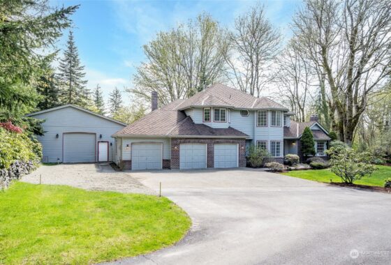 A two-story house with a three-car garage and driveway surrounded by trees under a blue sky.