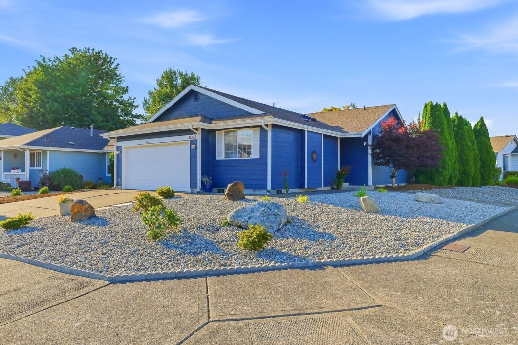 A single-story blue house with a white garage door sits on a gravel front yard under a clear blue sky.