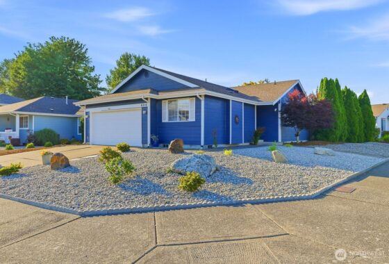 A single-story blue house with a white garage door sits on a gravel front yard under a clear blue sky.