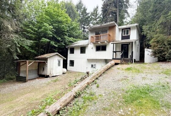 A white house with a wooden deck and porch surrounded by trees, featuring a small shed and gravel driveway.
