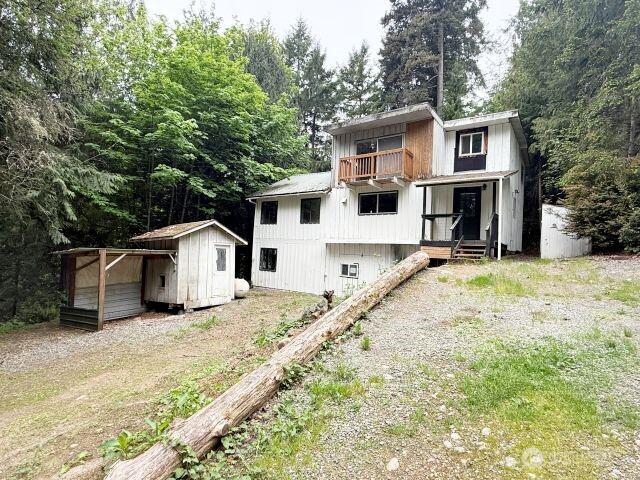 A white house with a wooden deck and porch surrounded by trees, featuring a small shed and gravel driveway.