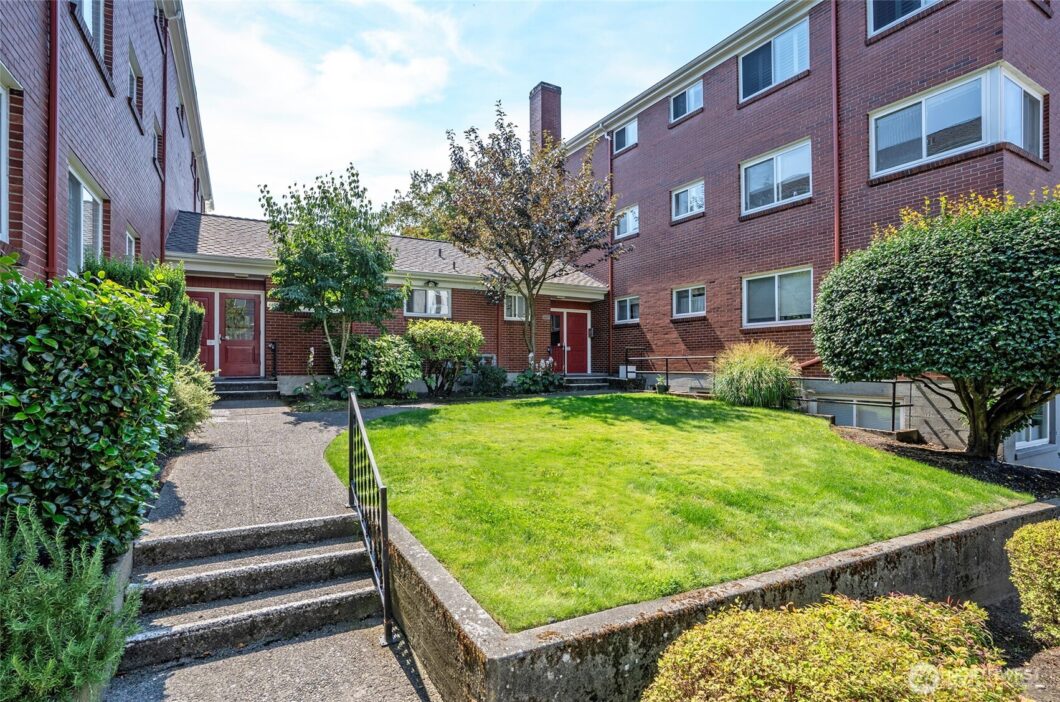 A brick apartment building with a red door and windows overlooks a green lawn, surrounded by trees and bushes under a blue...